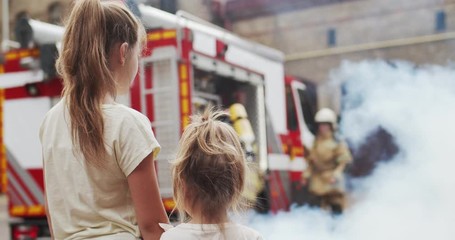 Portrait of two girls' sisters watching firemen fight fire. Firefighters in helmets and protective suits in the smoke saving people. The concept of saving lives, heroic profession, fire safety