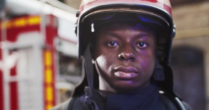 Portrait of African American firefighter approaches from fire van with flashing lights on and standing in front of camera looking. Concept of saving lives, heroic profession, fire safety
