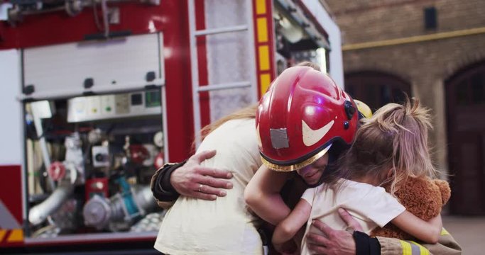 Middle plan injured little girls sisters reunites with their loving father fireman in helmet and gull equipment. In background fire truck. The concept of saving lives, heroic profession, fire safety