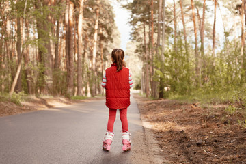 Back view of little charming female wearing red casual jacket and leggins, riding rolling skates,...