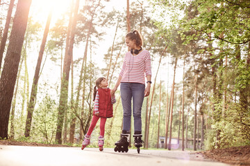 Mother and daughter riding on roller skates by road in forest, mommy looking at her cute girl,...
