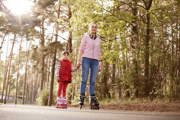 Mother with daughter roller skating in park together, family wearing casual outfits posing on road surrounded with trees, enjoying spending time in open air. - obrazy, fototapety, plakaty