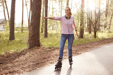 Happy young girl wearing casual striped shirt and jeans, roller skating in park, enjoying beautiful...