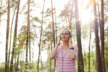 Adorable female wearing striped shirt listening to music in forest, woman posing surrounded with trees, touching her headphones with palms, enjoying music and nature.