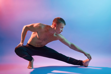 Man performing yoga, doing exercising for stretching, topless guy practicing yoga isolated over neon background, muscular male dresses black pants.