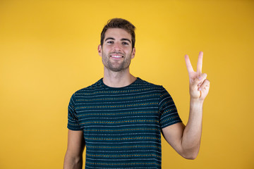 Fototapeta premium Young handsome man wearing casual blue t-shirt over yellow isolated background smiling with happy face winking at the camera doing victory sign. Number two.