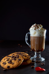 Close up of a glass cup of coffee with whipped cream and chocolate on it, chocolate chip cookies and roasted coffee beans on dark background. Concept of ready to eat food, tasty snack. Selective focus