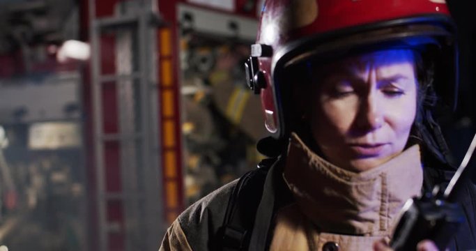 Portrait of upset scared on panic female fireman in helmet and gull equipment standing next to car with flash lights on at night screaming dispatcher on walkie-talkie and crying.