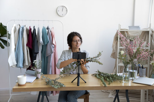 Smiling attractive young female professional decorator blogger sitting at table in front of mobile phone on stabilizer, filming educational diy bouquet video, recording online workshop in office.
