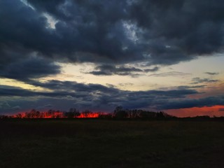 A panorama of a brilliant sunset sky between gray clouds and silhouettes of dark hills, with the last bit of sun slipping below the horizon.