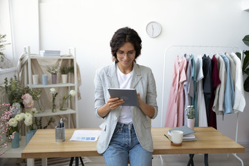 Smiling young attractive professional decorator leaning at table, communicating with clients on computer tablet indoors or using applications for creating sketching new clothing collection in office.