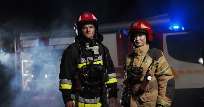 Middle plan of Caucasian man and woman firefighters in the full equipped costumes and helmets standing outside looking into camera at night in smoke next to fire van with flash lights on.