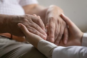Close up kind caregiver medical worker holding hands of old 70s retired patient, supporting at...