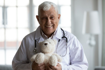 Head shot portrait of happy kind elderly mature skilled pediatrician doctor in medical uniform, holding fluffy toy in hands, waiting for small patients in modern clinic, healthcare service concept.