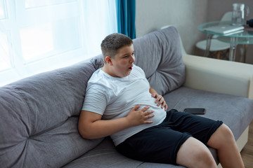fat teenager boy has stomach ache after junk food, sit on sofa holding belly, suffer from pain