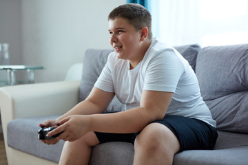 fat caucasian teenager boy enjoys video games, overweight boy sits on sofa holding console in...