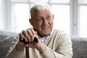 Head shot smiling old disabled man holding hands on wooden stick, resting on cozy couch in living...