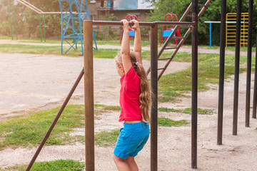 Girl hanging on horizontal bar. Child doing motion exercises and playing on playground outdoor. Healthy leisure sport activity on fresh air. Happy childhood. Kids physical training on sports stadium.