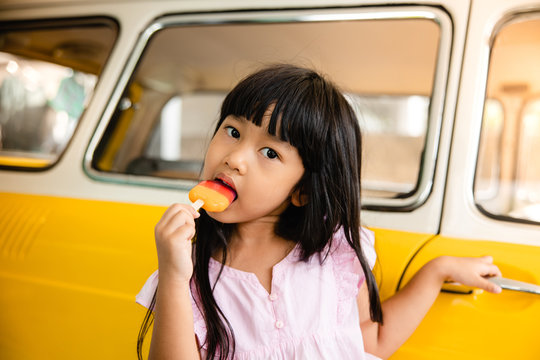 Asian Kids Eating Ice Cream Against A Yellow Car In Summer. Looking At Camera