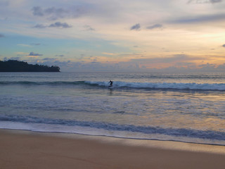 Fototapeta na wymiar A lone surfer rides on a wave in the evening at sunset. Deserted beach, clean sand. The glow of the sundown on a cloudy sky, twilight at the resort. A green hill in the distance stretches into the sea