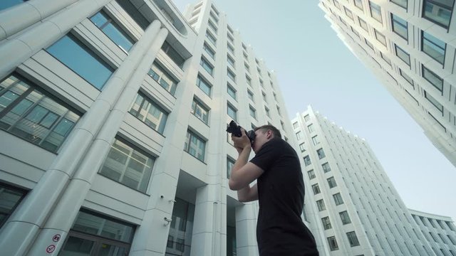Young photographer takes picture of modern urban architecture. Action. Bottom view of guy photographing modern architecture with camera. Photographer shoots office buildings against blue sky