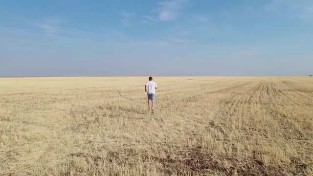 Athletic man runs across dried mown field after harvesting wheat into the distance, drone shooting from different angles. Feeling lonely or free, trying to get away from problems.