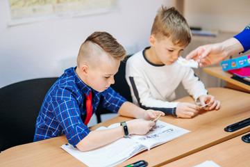 Concentrated schoolboys sitting at desk and writing in exercise book with classmate sitting behind.