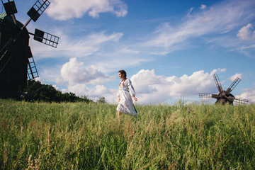 Young cheerful woman in traditional ukrainian long shirt have fun on countryside meadow. Happy beautiful girl in embroidery dress walking on summer field among mills. Carefree countryside lifestyle