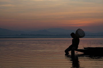 At lake, young asian man holding bamboo fish trap for catching fish and sitting on the boat in the morning