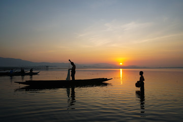 Fototapeta premium asian fisherman standing on boat using fishing net to catch fish while his son standing and holding jar in the lake at the sunrise