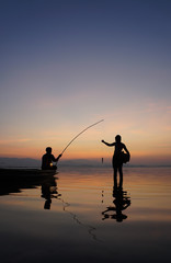 At lake side, asian fisherman sitting on boat while his son standing and  using fishing rod to catch fish at the sunrise