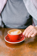 Woman's hand with red nails holding red cup of coffee on the table. Close up photo of coffee and woman's hand horizontal view. Woman wearing grey sweater.