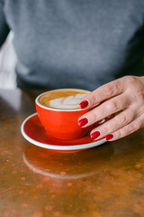 Woman's hand with red nails holding red cup of coffee on the table. Close up photo of coffee and woman's hand horizontal view. Lady wearing grey warm sweater.