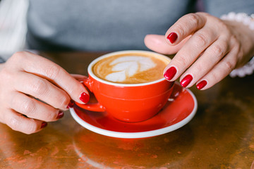 Woman's hands with red manicure holding red cup of coffee on the table. Close up photo of coffee art and woman's hand horizontal view. Woman wearing grey sweater.