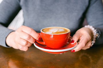 Woman's hand with red nails holding red cup of coffee on the table. Close up photo of coffee and woman's hand horizontal view. Business woman wearing grey sweater.