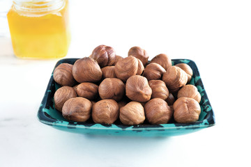 Raw peeled hazelnuts in a rectangular green bowl with a black pattern stands on a white background. In the background is a small jar of honey. Delicious and healthy dessert, snacks. Close-up