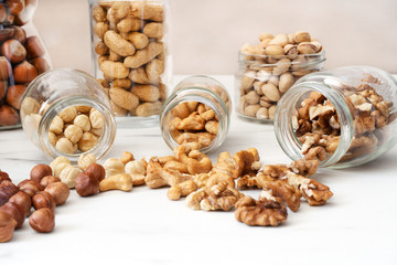 Three glass jars with nuts (peeled hazelnuts, cashews, walnuts) lie on a white table. Nuts spilled out of the jars. In the background are jars of pistachios, hazelnuts and shells