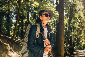 Portrait of mature hiker with backpack and stick standing in forest.  Senior man taking a break during trip.