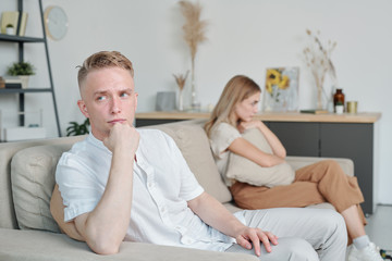 Young offended or annoyed man sitting on couch while ignoring his unhappy wife