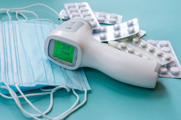 Surgace thermometer with digital green colored screen showing high body temperature on pile of medical masks and pills in blisters. Diagnostics body temperature