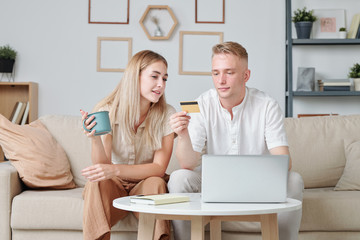 Contemporary young husband and wife looking at credit card in hands of man