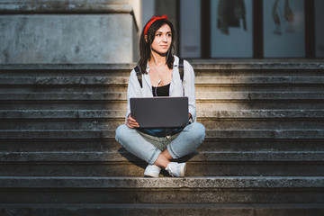 Beautiful happy relaxed sun-kissed young woman, working on laptop, casual contemplating while sitting on the university's stairs, thinking about going back to school