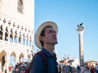 Young handsome man in San Marco square, Venice, Italy. Tourist with panama hat and backpack on the background of the Doge's Palace and Lion of St. Mark. Cultural and historical tour. Travel in Europe.
