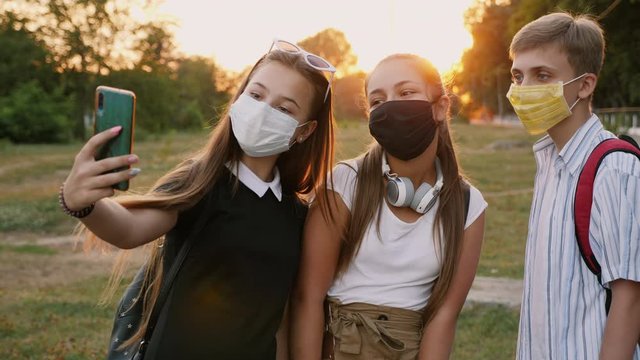 Teenagers with backpacks and wearing protective masks on their faces take a selfie on a smartphone in a city park at sunset