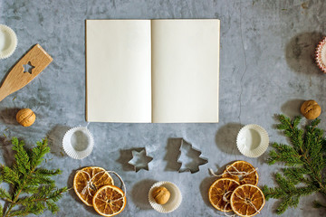 Christmas food recipe background. A book for writing recipes on a dark table surrounded by fir branches, dried oranges, paper molds for cupcakes, nuts. Top view, flat lay, copy space