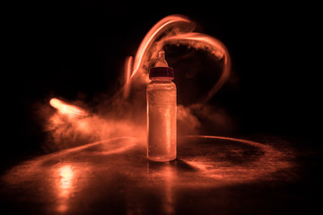 Close up of baby bottle at wooden table on dark toned foggy background with light. Creative artwork decorated photo.