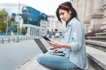 Portrait of a female student with headphones using laptop while sitting on steps near university