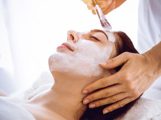 Beautiful brunette woman enjoying applying cosmetic mask with closed eyes in sunny spa center. Relaxing treatment and medicine concept