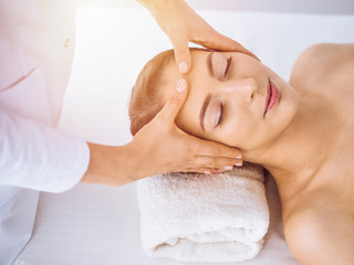 Fototapeta na wymiar Beautiful brunette woman enjoying facial massage with closed eyes in sunny spa center . Relaxing treatment and cosmetic medicine concepts