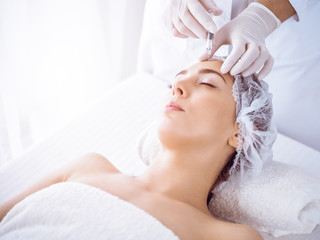 Obraz na płótnie Canvas Beautician doing beauty procedure with syringe to face of young brunette woman in sunny spa center. Cosmetic medicine and surgery, beauty injections